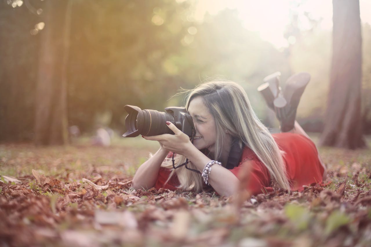 A young woman lies on the ground, capturing the beauty of fall through her camera in a serene park.